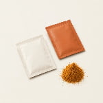 Two flat three-side seal sachets rest beside a small mound of warm-colored spice powder.