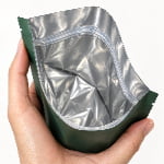 A hand holds open a green stand-up pouch with a metallic silver foil lining and thin clear press-to-close zipper tracks