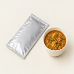 A flat silver retort pouch lies beside a simple bowl of ready-to-eat vegetable curry.